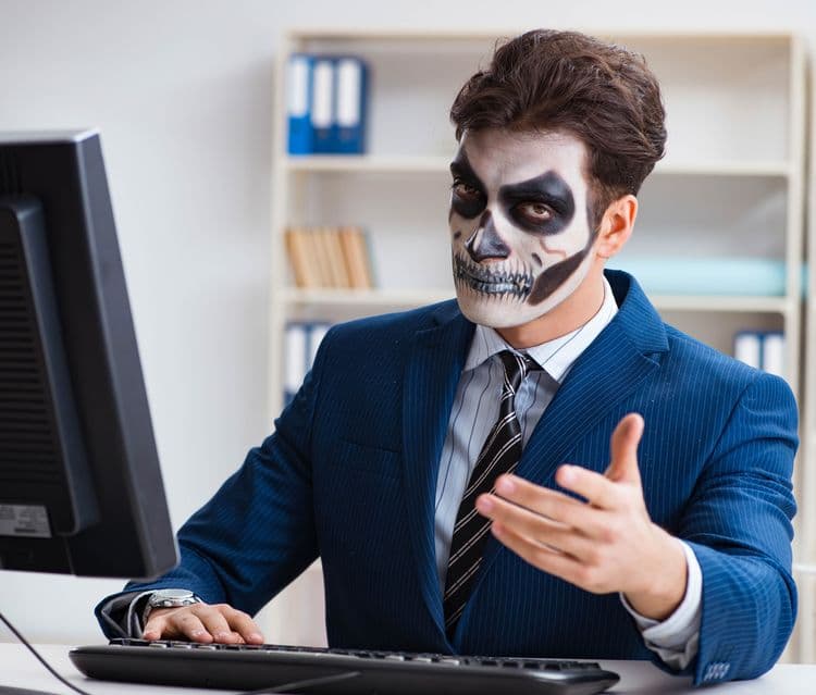 Executive in a suit with a frightening mask painted on his face, sitting at a computer and illustrating the charade of the annual performance review